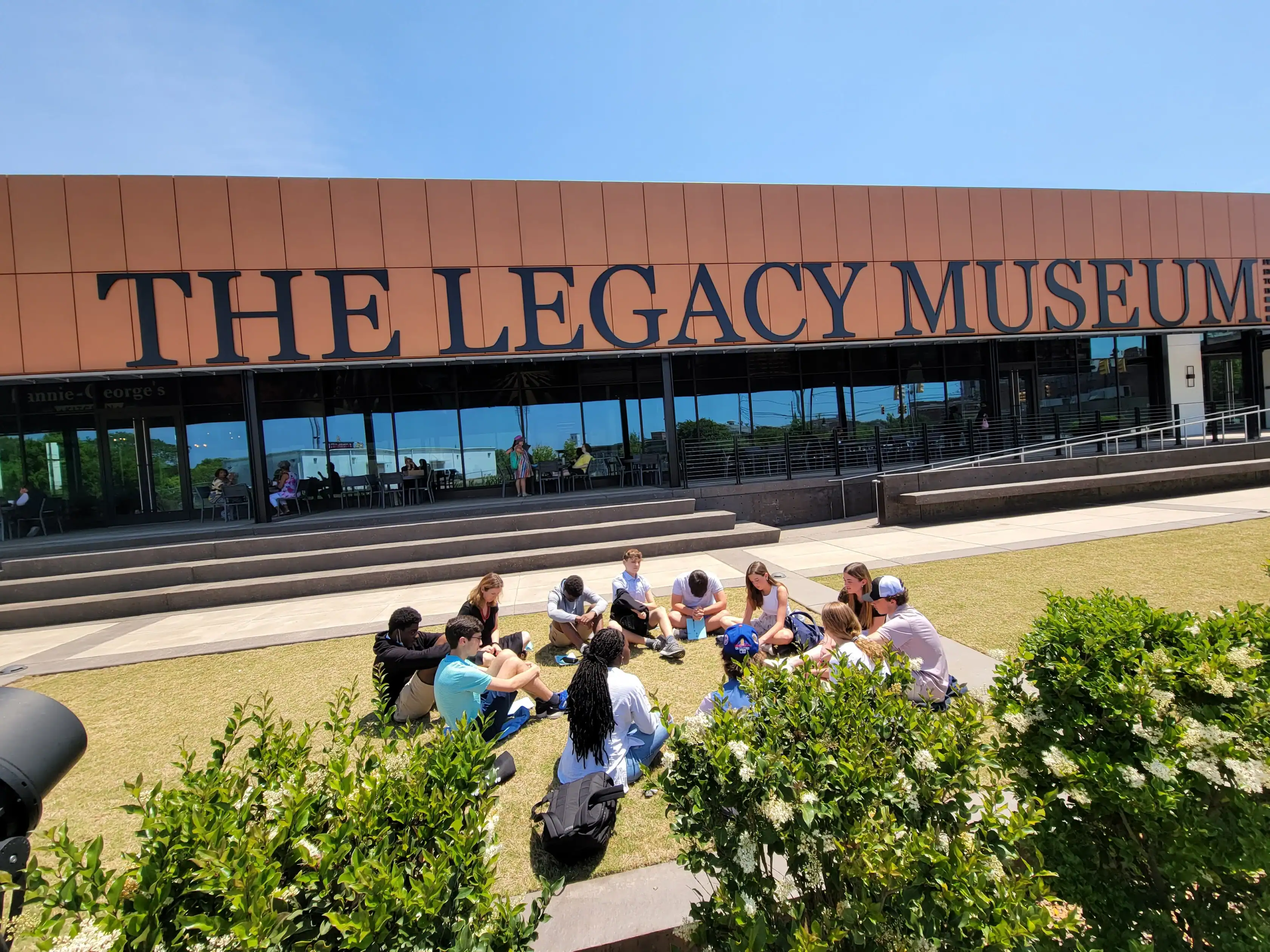 A group of people sits in a circle on the grass in front of The Legacy Museum building on a sunny day, enjoying a civil rights school trip.