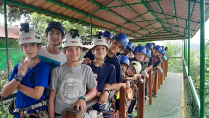 A group of people wearing helmets and harnesses stand on a covered platform, smiling at the camera. They're ready for a zip lining adventure.