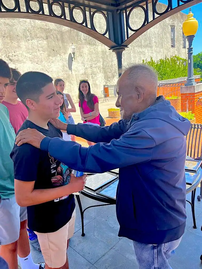 An older man places his hands on a teenage boy’s shoulders during a civil rights school trip, while others stand nearby under a metal canopy in an outdoor setting.