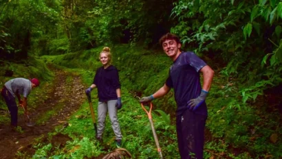 Three people work on a forest trail, holding shovels. Two are smiling and standing, while one bends forward, possibly digging. The area is lush with greenery.