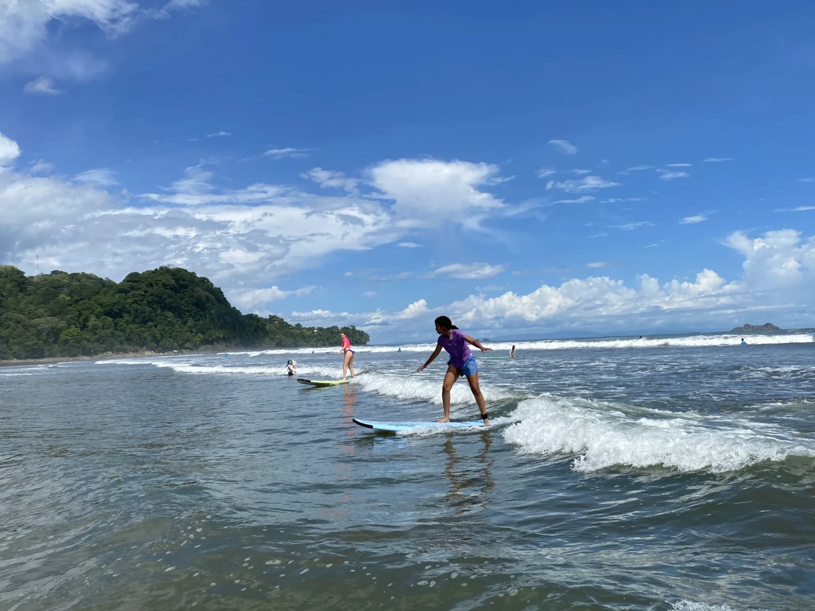 Teens on a medical trip abroad enjoy surfing small waves near the shore on a sunny day, with a forested hill and blue sky in the background.