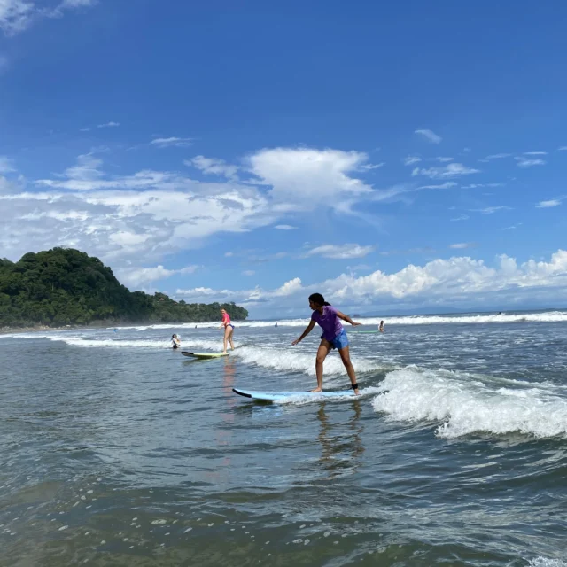 Teens on a medical trip abroad enjoy surfing small waves near the shore on a sunny day, with a forested hill and blue sky in the background.