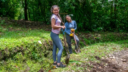 Two people smiling while digging in a garden with shovels, surrounded by lush greenery.