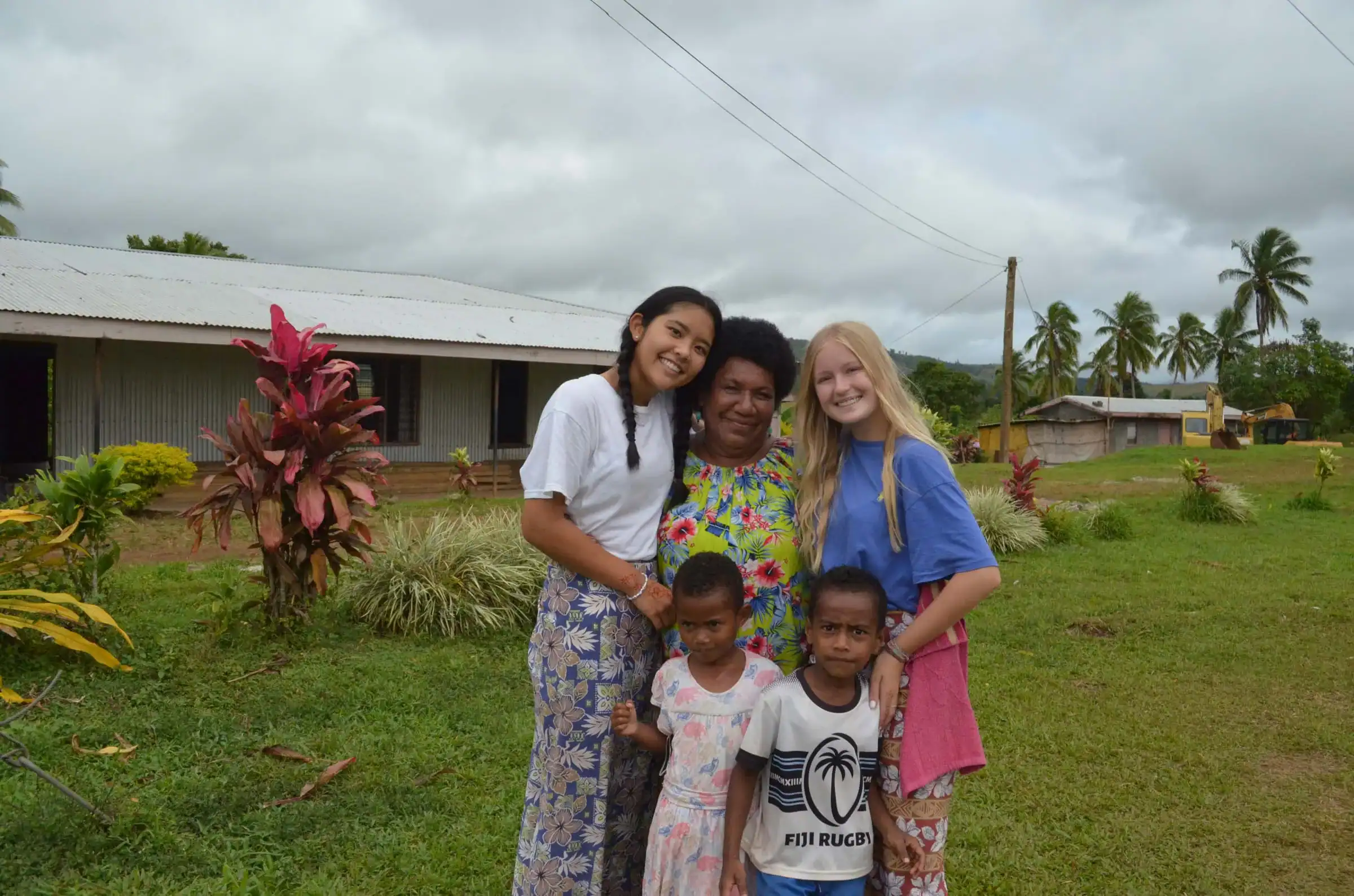 Five people, including three adults and two children, stand together outside on grass with buildings and palm trees in the background, smiling at the camera during a Fiji student trip.