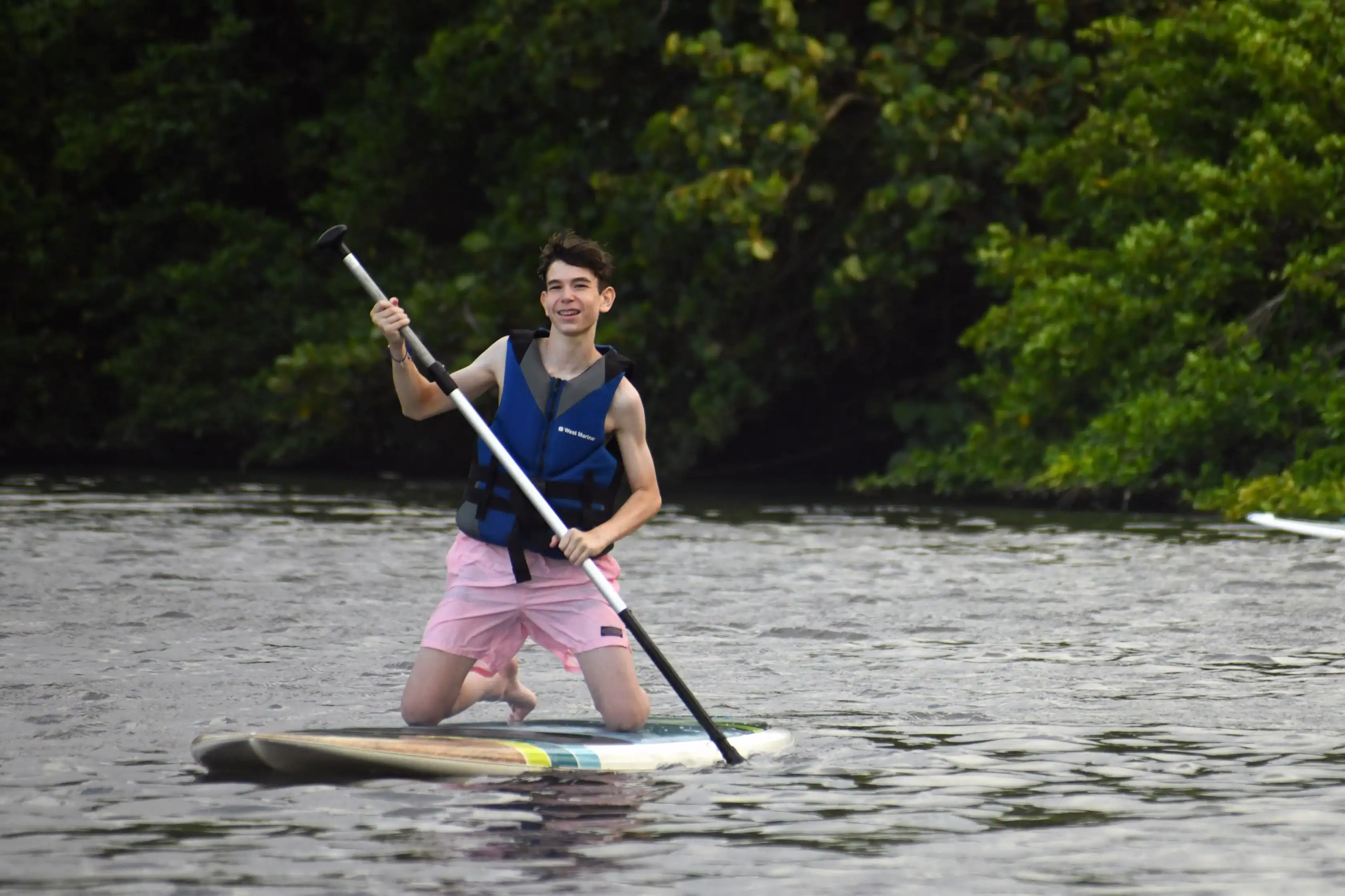 A person on a Puerto Rico service trip for teens wears a blue life jacket and pink shorts while kneeling on a paddleboard, holding a paddle on a river with trees in the background.