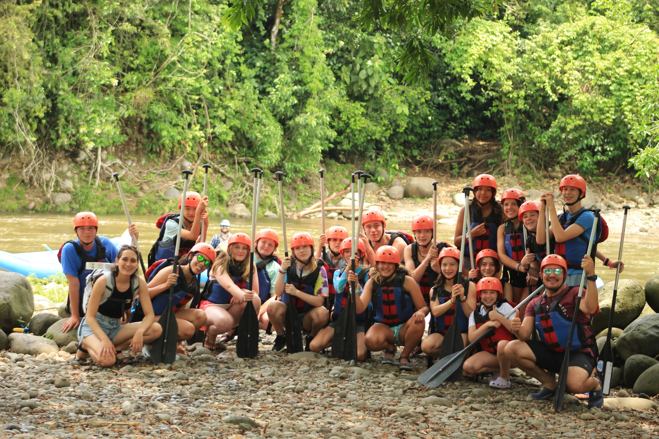 A group of people wearing life jackets and helmets pose with paddles on a rocky riverbank surrounded by greenery, enjoying an adventurous outing as part of an environmental school travel program.