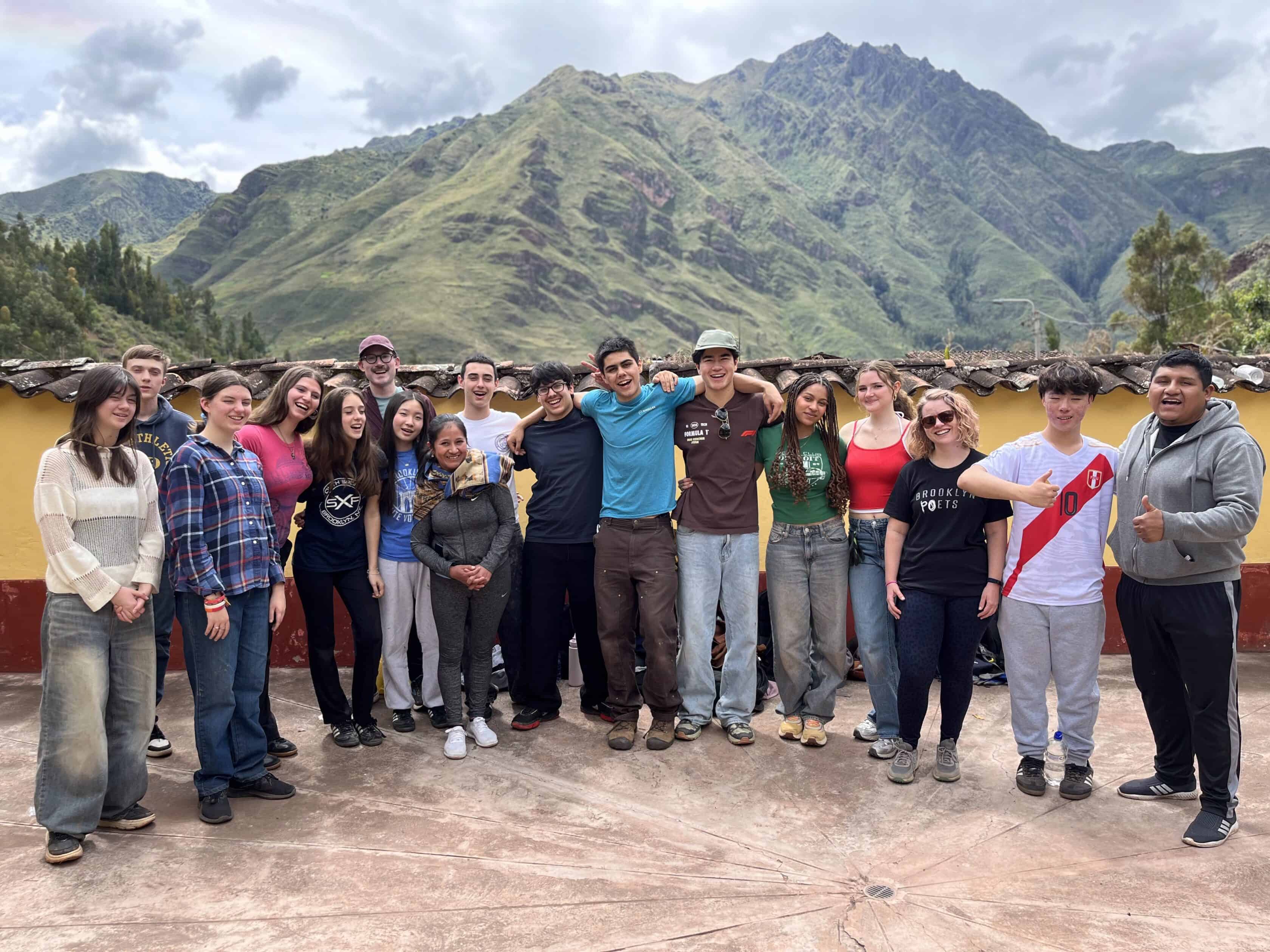 A group of students, part of a Peru student travel program, posing together outdoors on a terrace with mountains and a cloudy sky in the background.