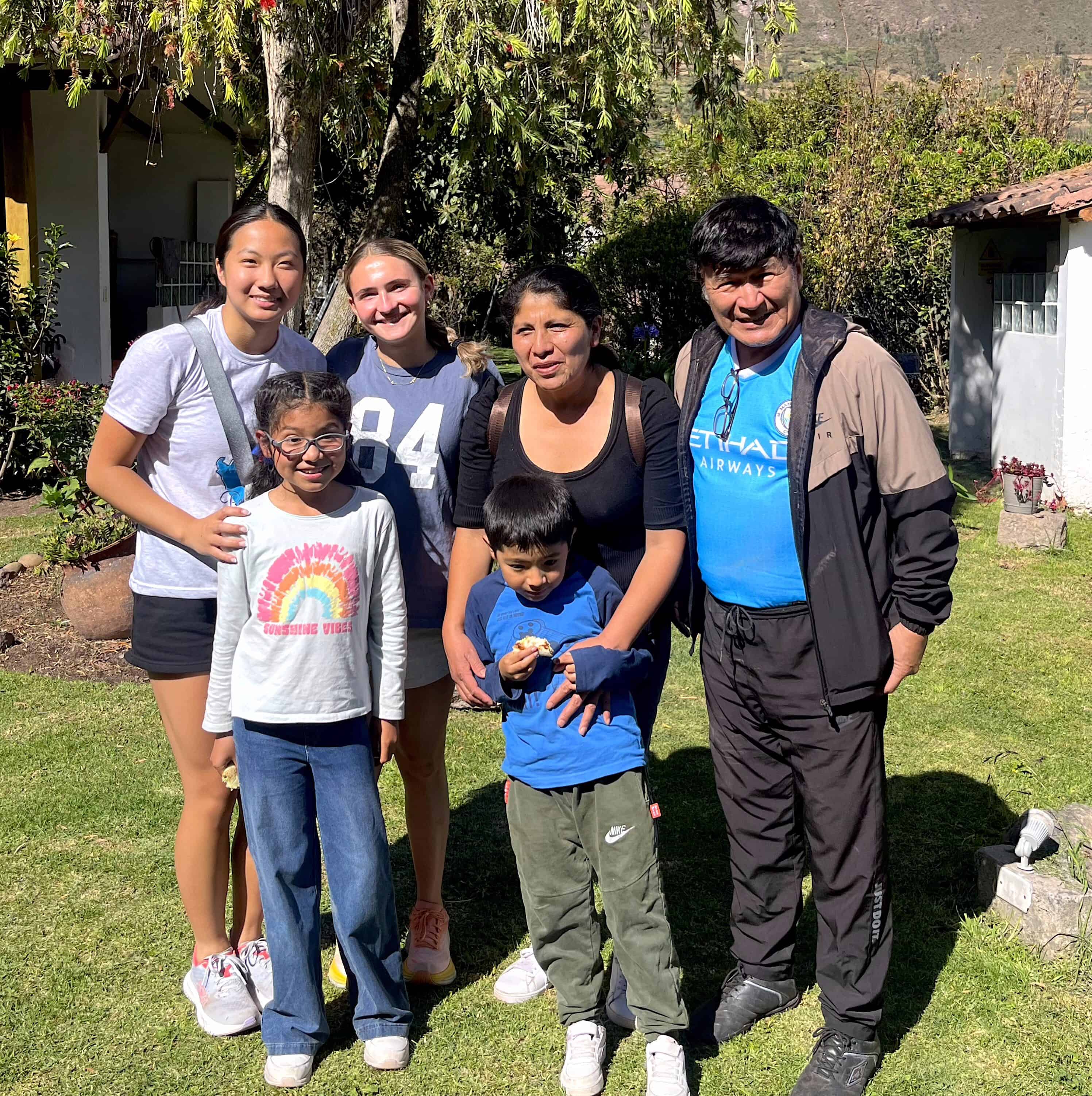 Six people—four adults and two children—stand together outdoors on a sunny Peru student travel adventure, smiling at the camera with greenery and buildings in the background.