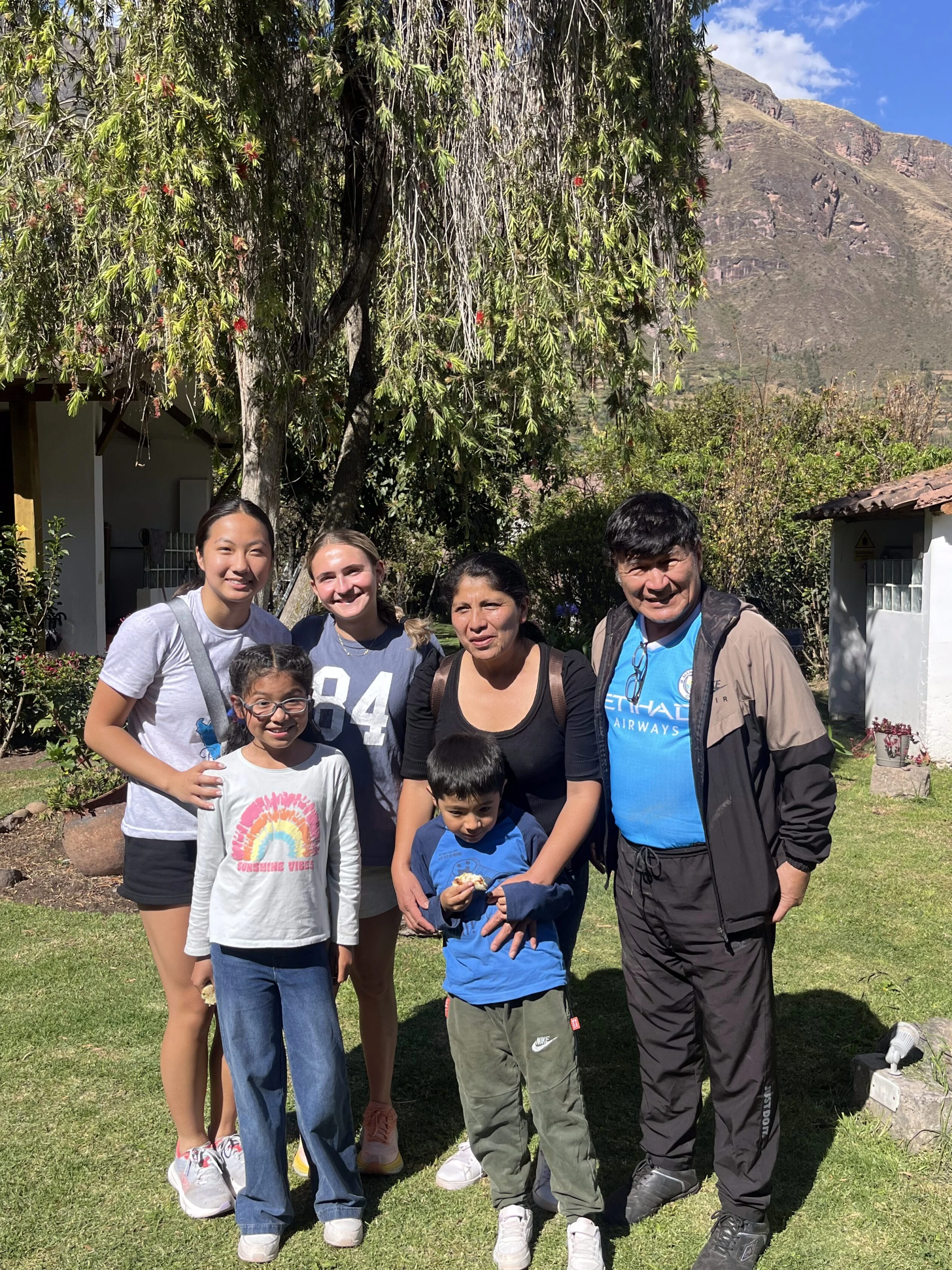 Six people, including two children and four adults, stand together and pose for a photo outside on a sunny day with trees and mountains in the background, capturing memories from their teen travel program.