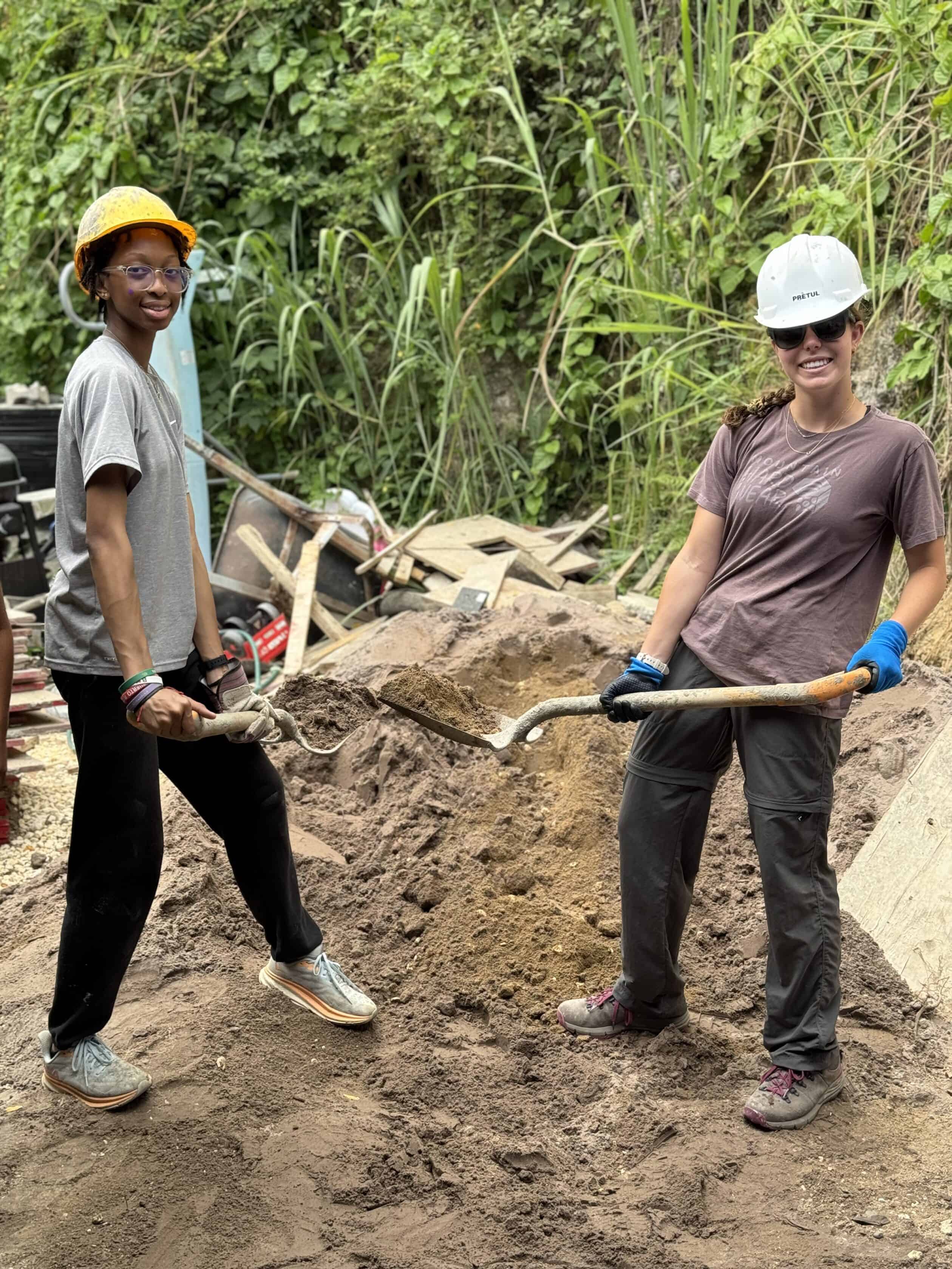 Two people on a teen service trip for teens in Puerto Rico wear hard hats and gloves as they stand on dirt, holding shovels and smiling, with construction materials and greenery in the background.