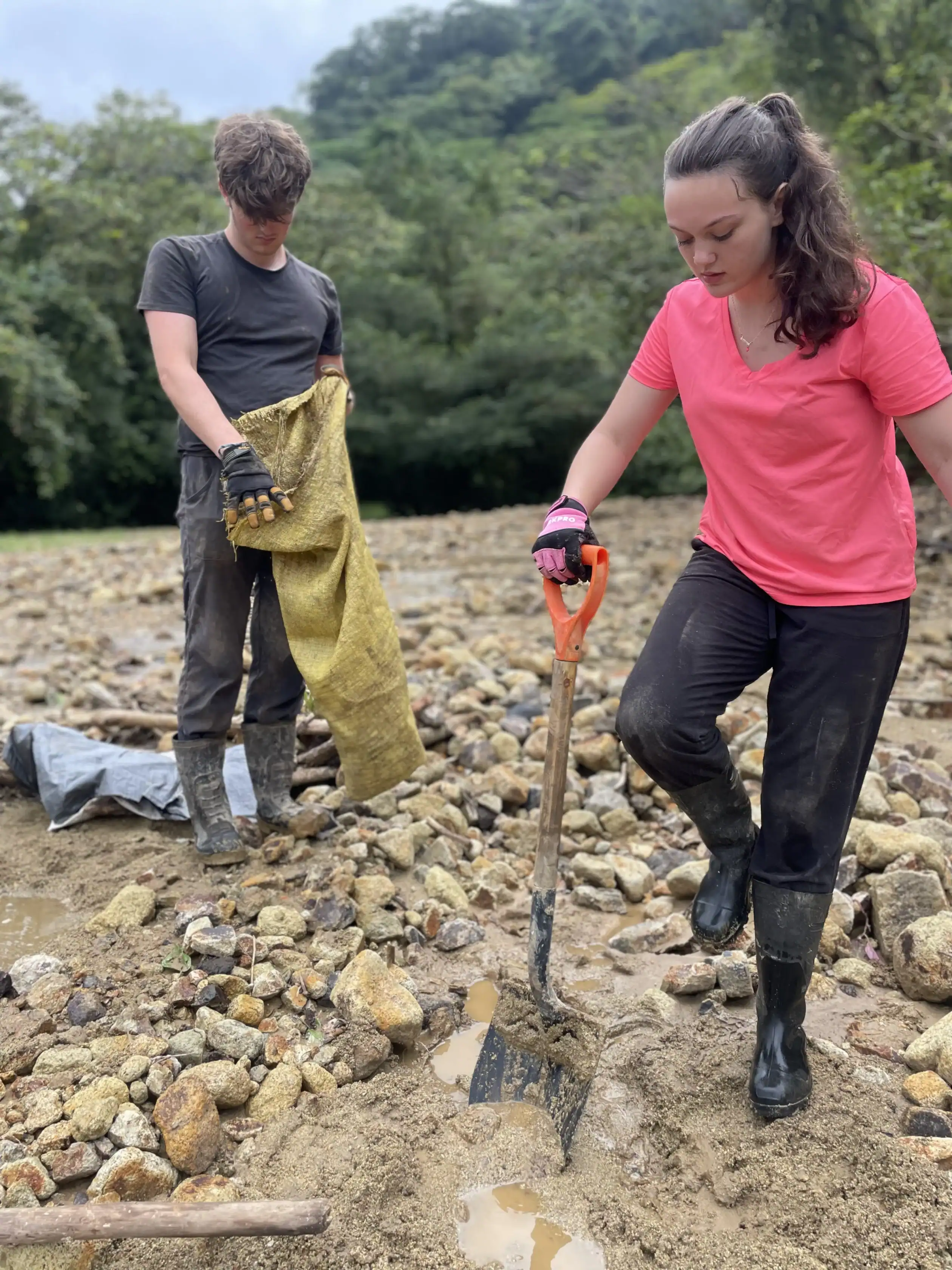 Two people on a high school science and conservation trip work outdoors on rocky ground; one lifts a shovel into the dirt while the other holds a yellow sack. Both wear gloves and boots, with trees visible in the background.