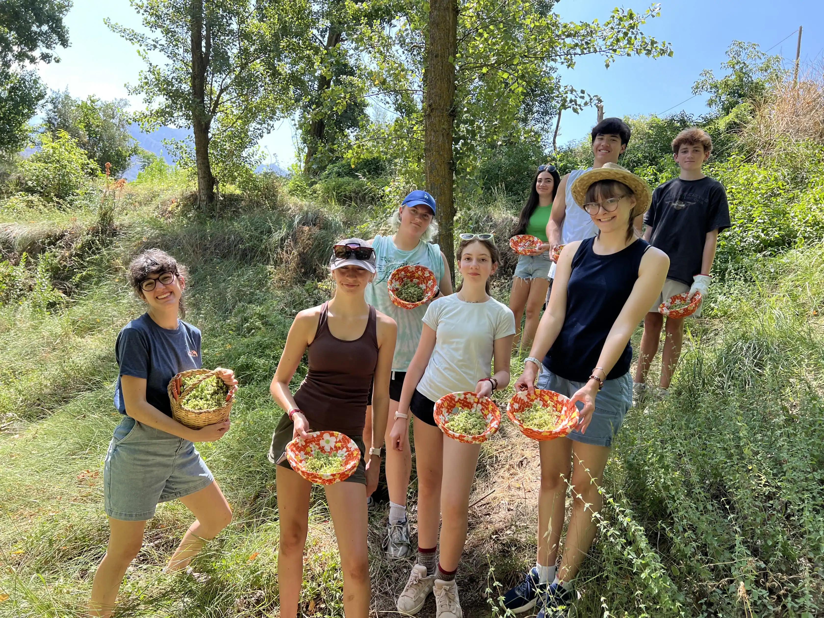 A group of eight young people stand outdoors in a grassy, wooded area, each holding a basket of freshly picked produce during a Spain summer travel program.