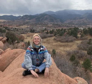 A person sits on a rocky outcrop wearing a colorful jacket, with a mountainous landscape in the background under a cloudy sky.