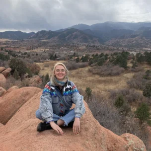 A person sits on a rocky outcrop wearing a colorful jacket, with a mountainous landscape in the background under a cloudy sky.