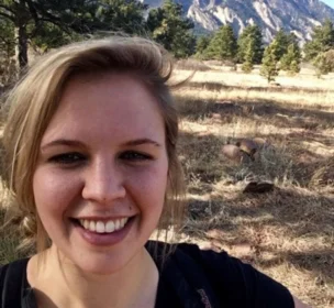 A person smiling in a grassy field with trees and mountains in the background.