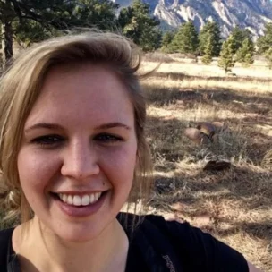 A person smiling in a grassy field with trees and mountains in the background.