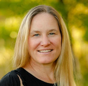 Woman with long blonde hair smiling outdoors with blurred green background.