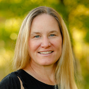 Woman with long blonde hair smiling outdoors with blurred green background.