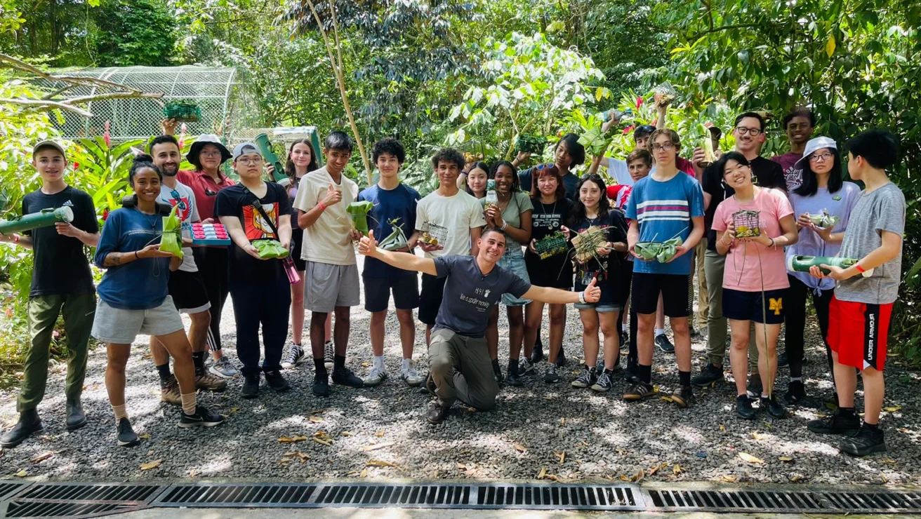 A group of people of various ages stand outside on a gravel path in lush greenery, holding plants and smiling at the camera.