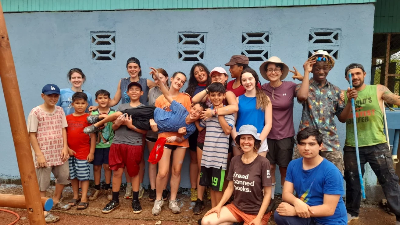 A diverse group of people, including children and adults, pose happily in front of a blue wall outdoors. They are dressed in casual summer clothes.