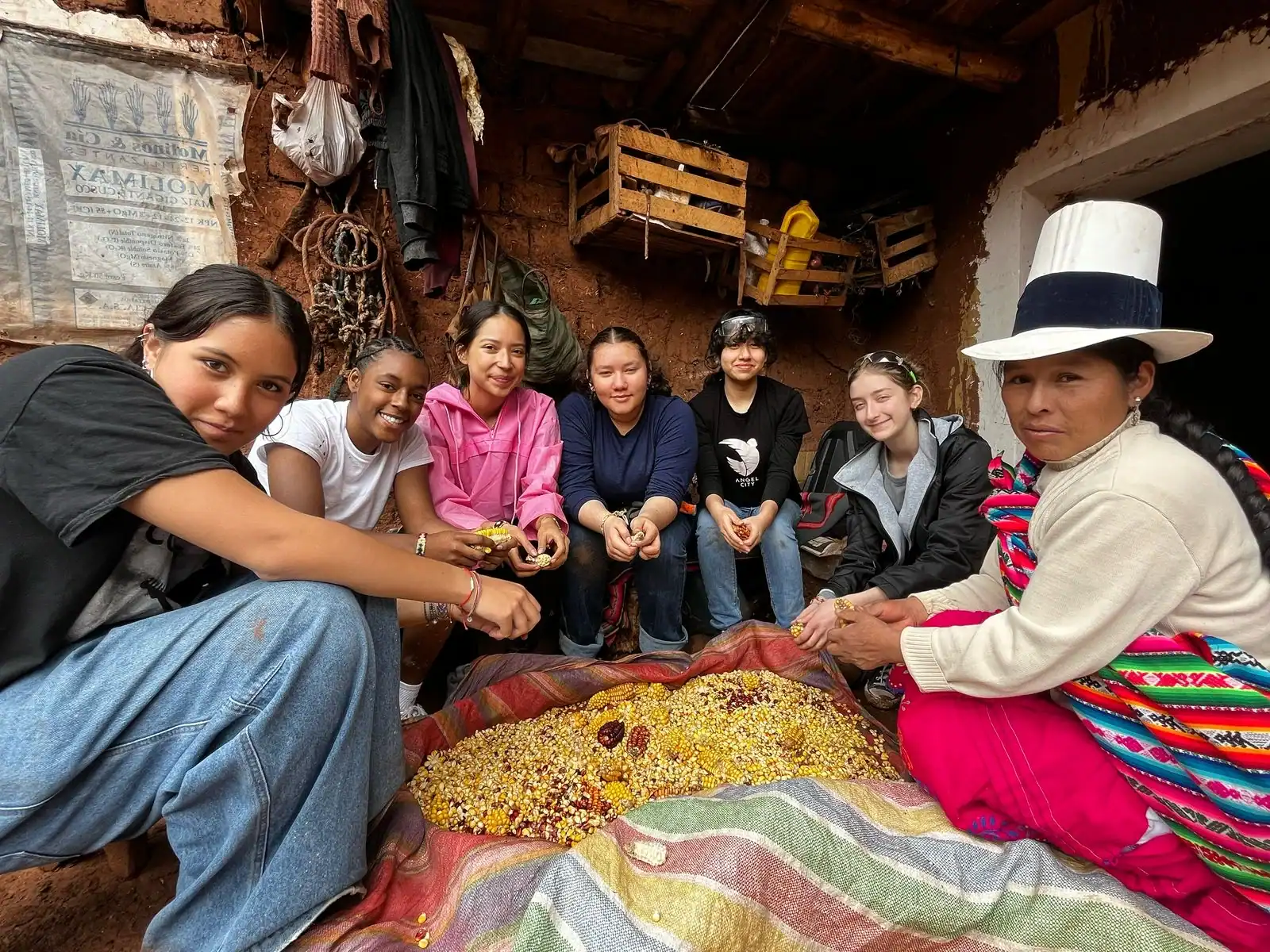 Students sit around a colorful pile of corn kernels inside a rustic building, with baskets and tools visible in the background—a scene that offers an authentic glimpse into local life for those on a Peru student travel experience.