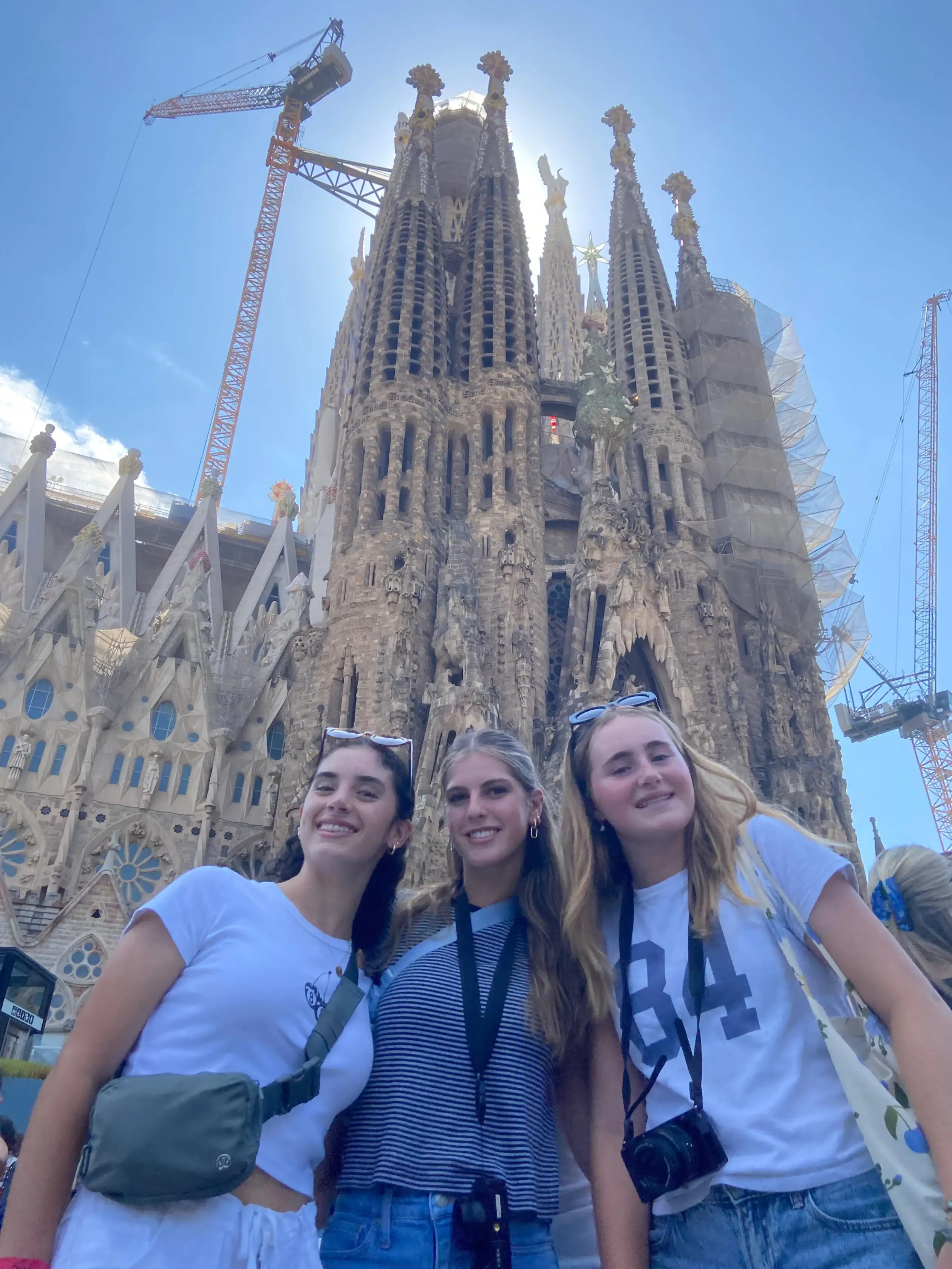Three young women stand smiling in front of the Sagrada Familia basilica in Barcelona, enjoying a sunny day as part of a Spain summer travel program, with cranes visible and a clear blue sky overhead.