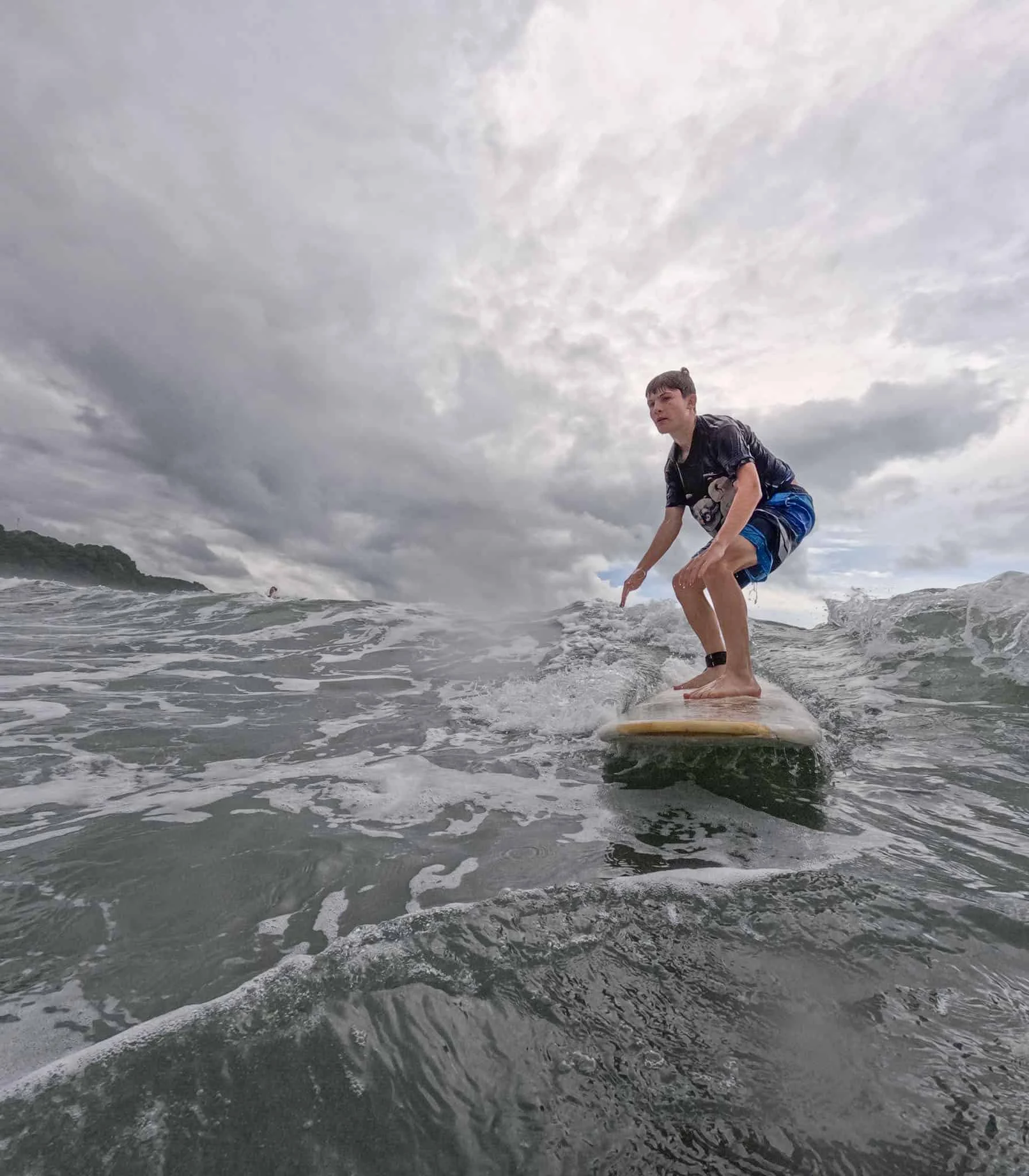 A teen enjoys a summer trip, riding a wave on a surfboard under a cloudy sky, with the ocean and distant shoreline visible.