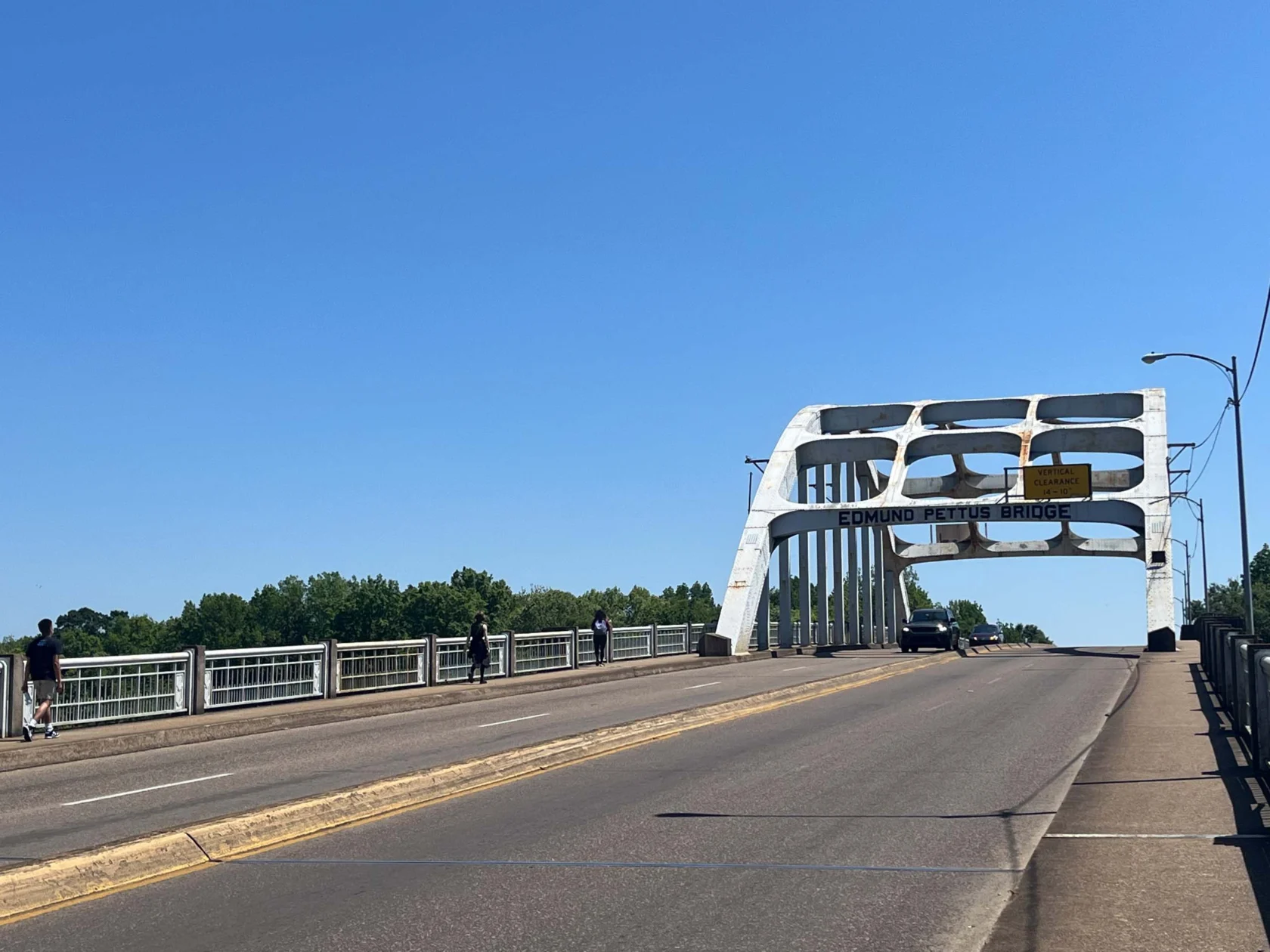 Edmund Pettus Bridge spans a clear blue sky with pedestrians on the walkway and a car driving across.