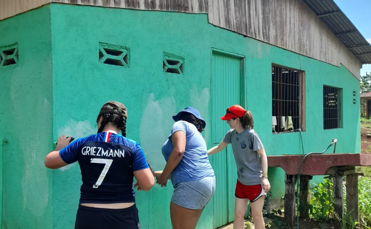 Three people repaint a green exterior wall of a building on stilts under a clear sky.
