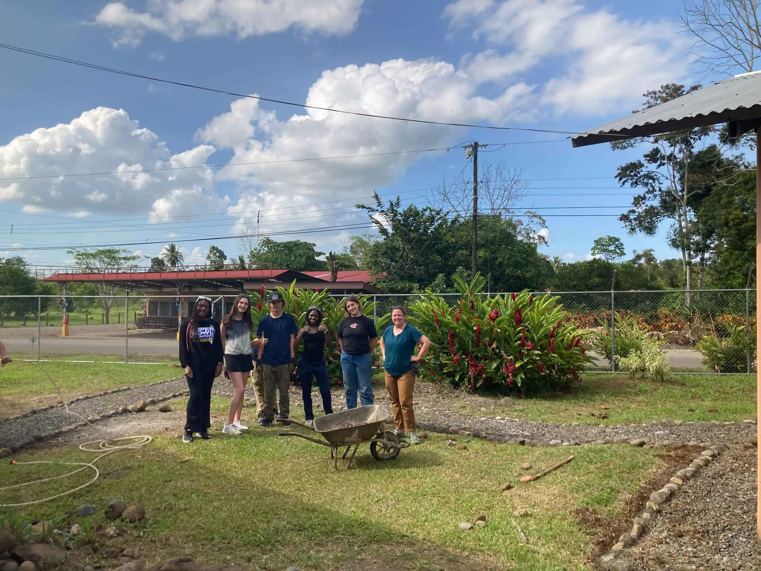 A group of six people stands outdoors on grass near a wheelbarrow, participating in an environmental school travel program, with greenery, a fence, and clouds in the background.