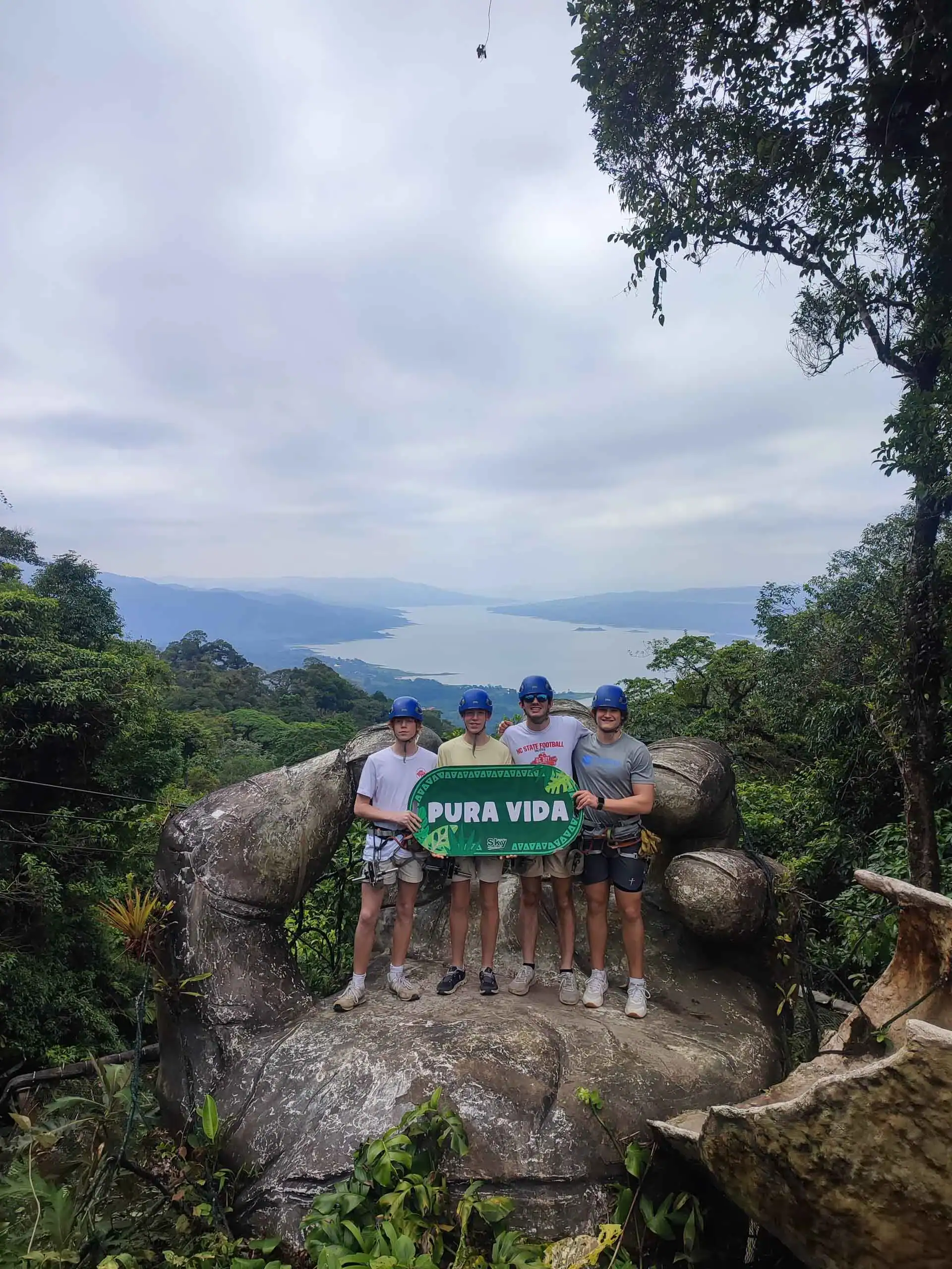 Four people wearing helmets stand on a large hand-shaped stone structure, holding a "Pura Vida" sign—a perfect moment from their environmental school travel program, surrounded by lush greenery and a scenic lake in the background.