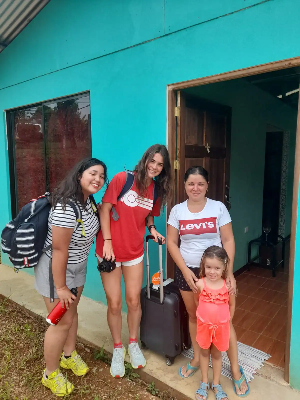 Four people, including a child, stand smiling outside the entrance of a turquoise house; one woman holds a suitcase and camera, ready for their Spanish immersion school trip.