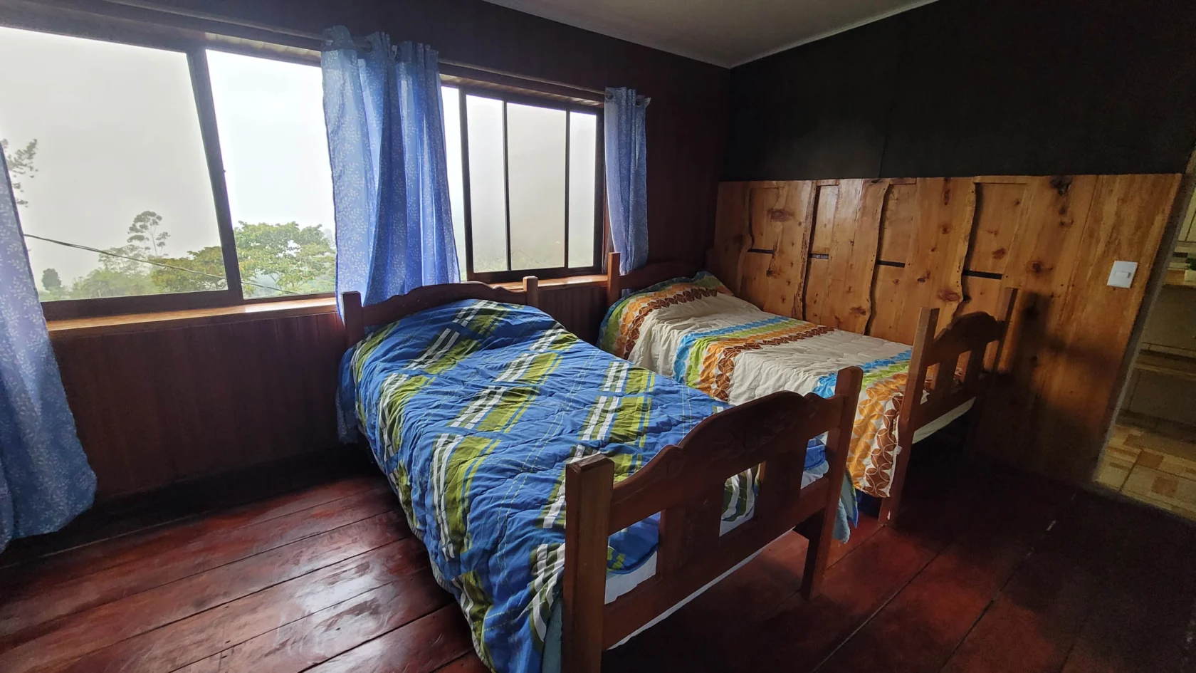 A small wooden bedroom with two single beds, each covered with different blankets. A large window with blue curtains overlooks greenery outside.