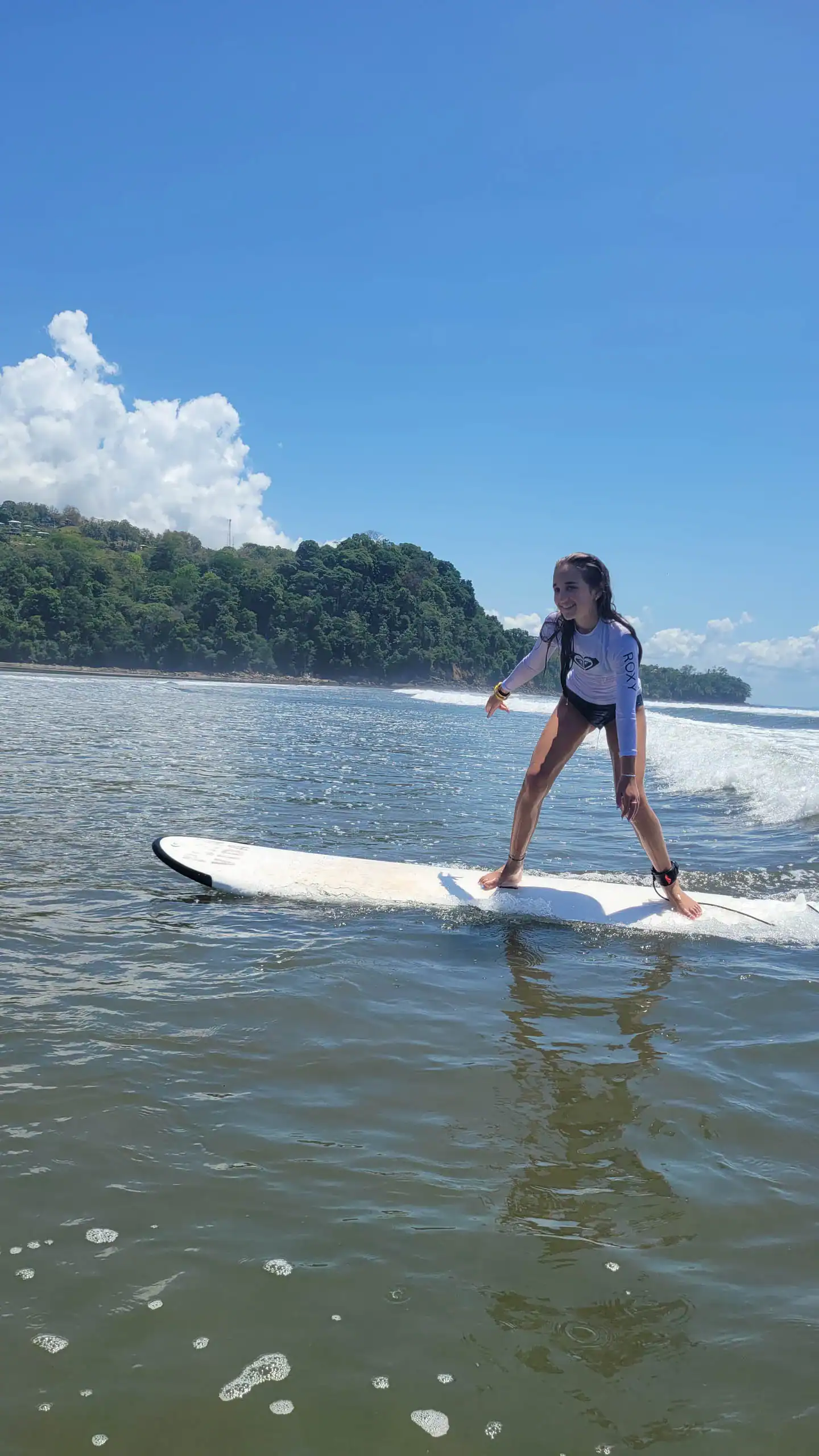 A person rides a surfboard on calm ocean water near a forested coastline under a blue sky with some clouds during a Spanish immersion school trip.