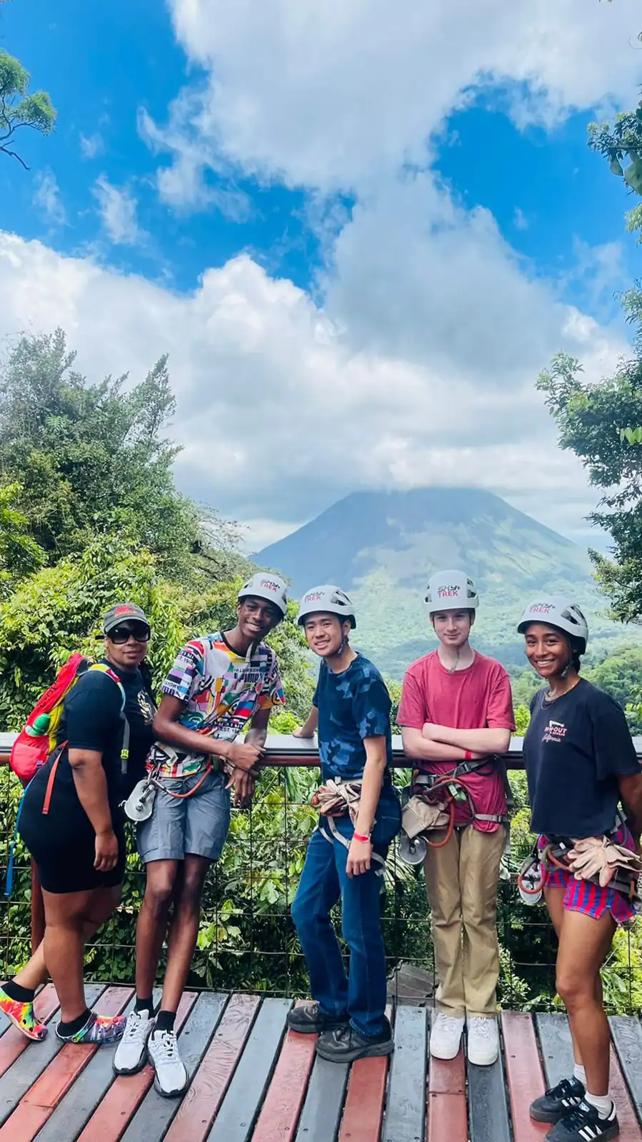 Five people wearing helmets and harnesses stand on a platform during a Spanish immersion school trip, with greenery and a distant volcano under a cloudy sky in the background.