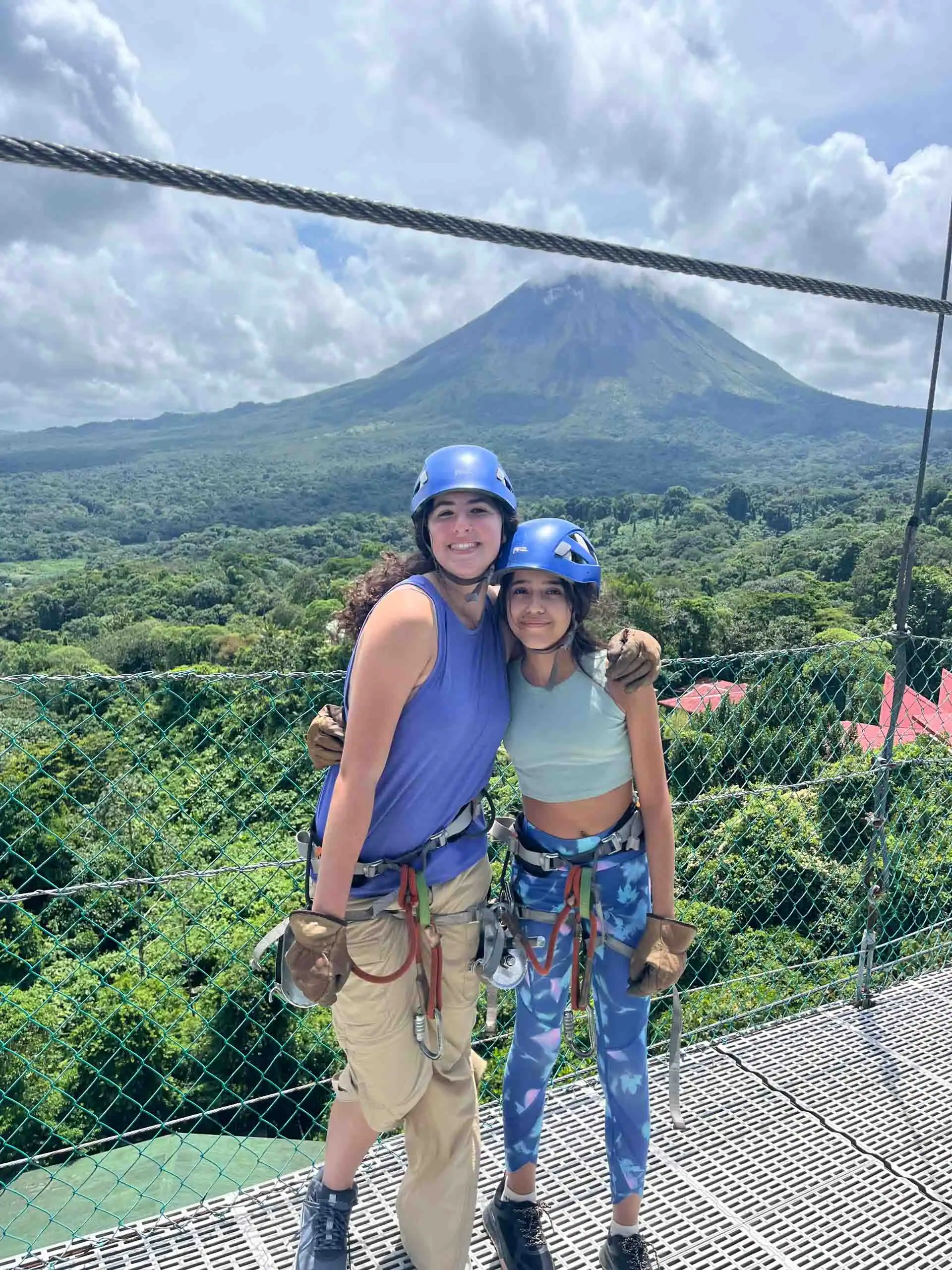 Two people wearing helmets and harnesses stand on a platform, ready for adventure as part of a Costa Rica Travel Program, with a lush green landscape and a large volcano in the background.