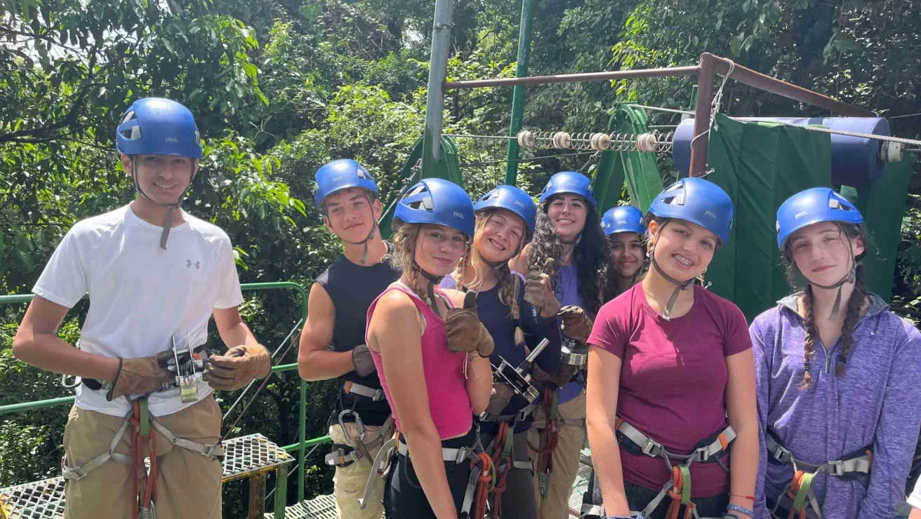 A group of people wearing helmets and harnesses, standing on a platform in a forest setting, preparing for a zip-lining activity.