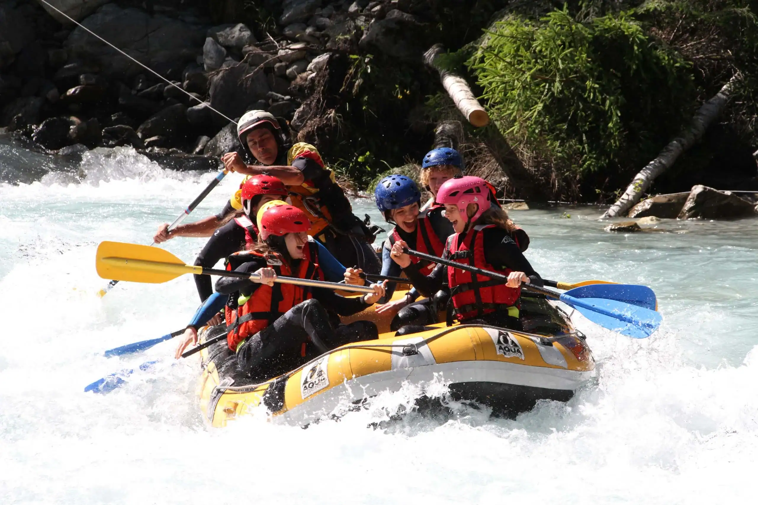 A group of people wearing helmets and life jackets are whitewater rafting on a fast-flowing river, paddling energetically through the rapids.
