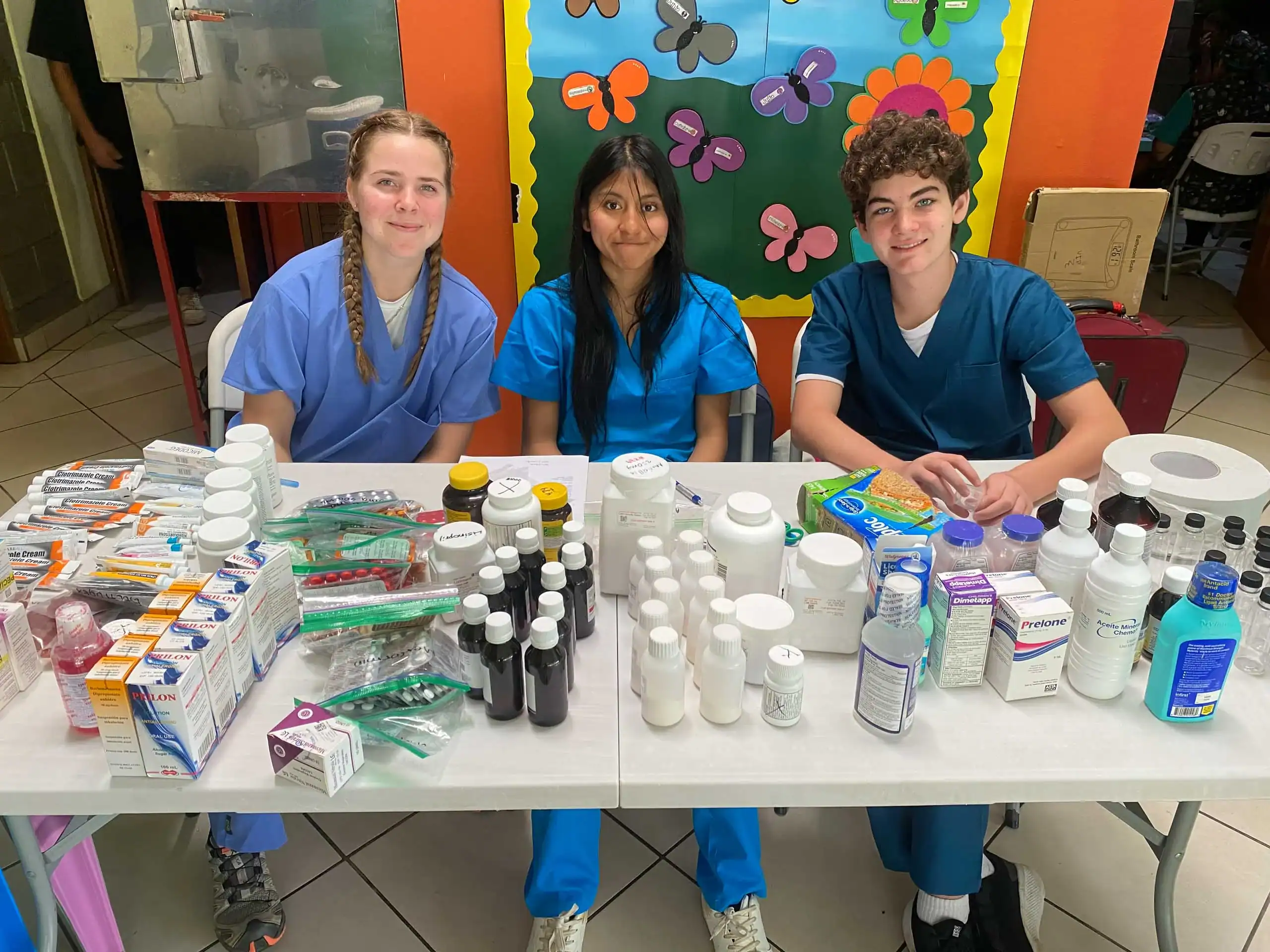 Three young people in scrubs sit behind a table filled with medical supplies and medications, in a brightly colored room with butterfly-themed wall decor, ready to share their expertise during a Dominican Republic school trip.