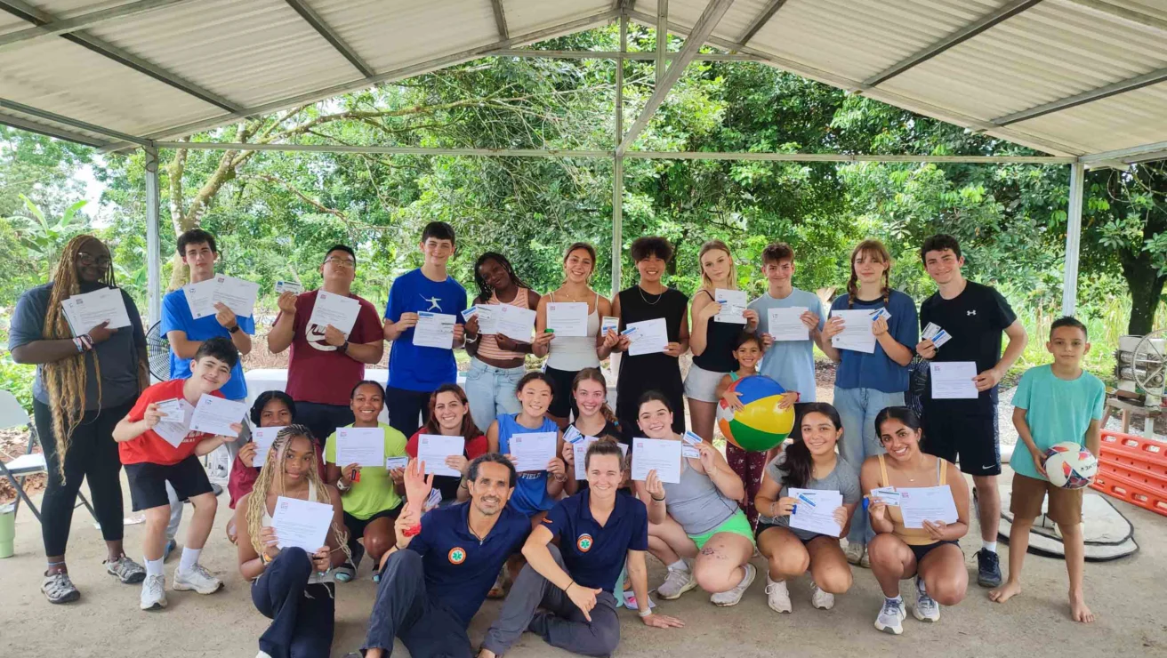 A group of people under a pavilion holding certificates and smiling. Some are holding a volleyball. Trees are visible in the background.