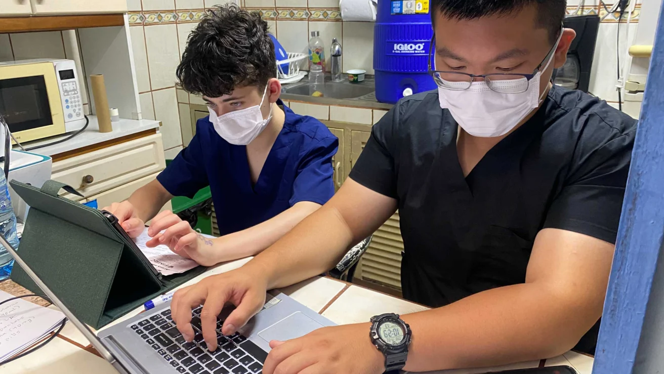 Two people wearing face masks work on a laptop and papers at a kitchen table, with a microwave and water cooler in the background.