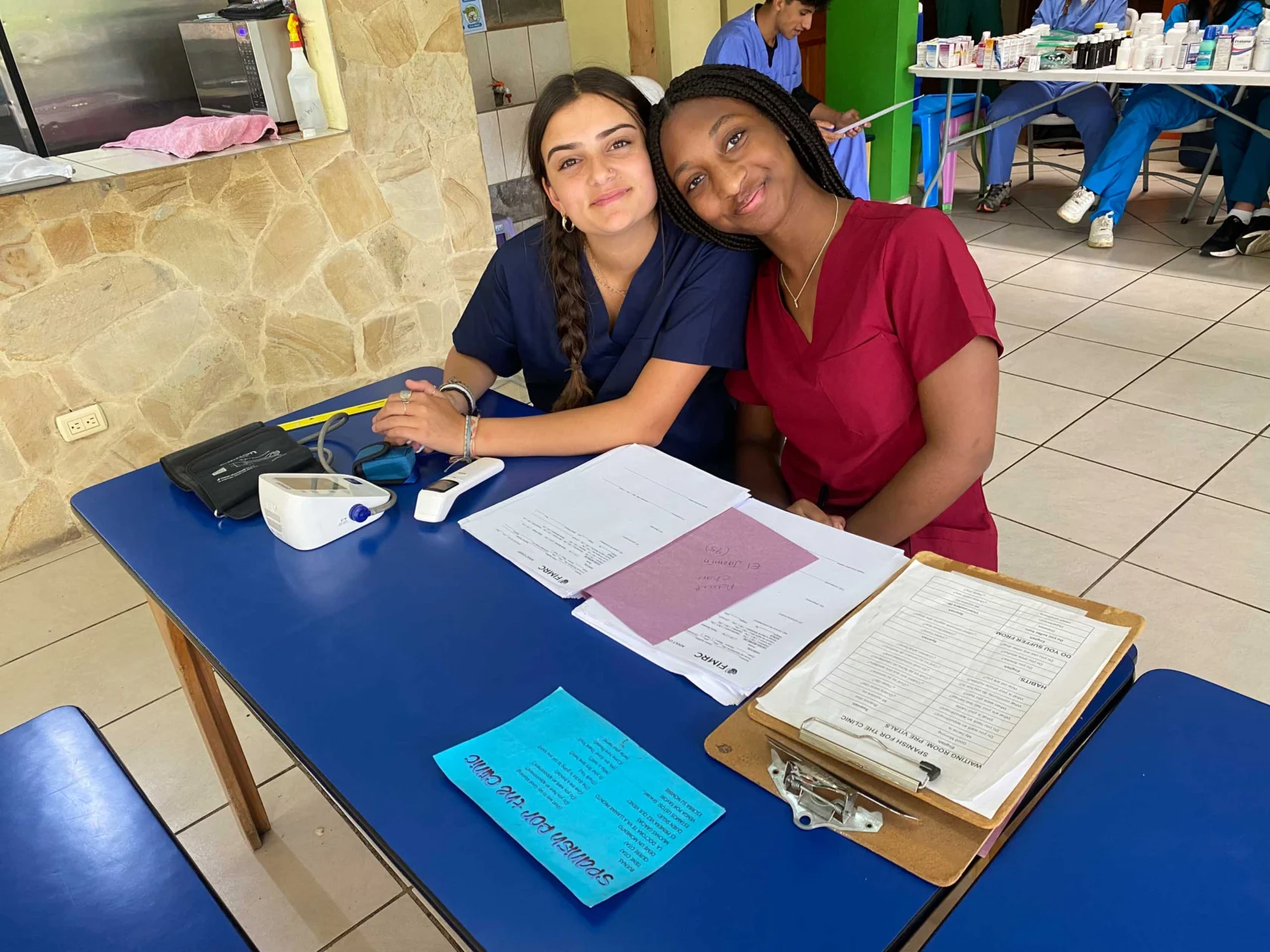 Two women in scrubs sit at a blue table with medical forms, a blood pressure monitor, and clipboards, smiling at the camera in a clinic during a medical study abroad experience in Costa Rica.