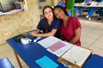 Two women in scrubs sit at a blue table with medical forms, a blood pressure monitor, and clipboards, smiling at the camera in a clinic during a medical study abroad experience in Costa Rica.