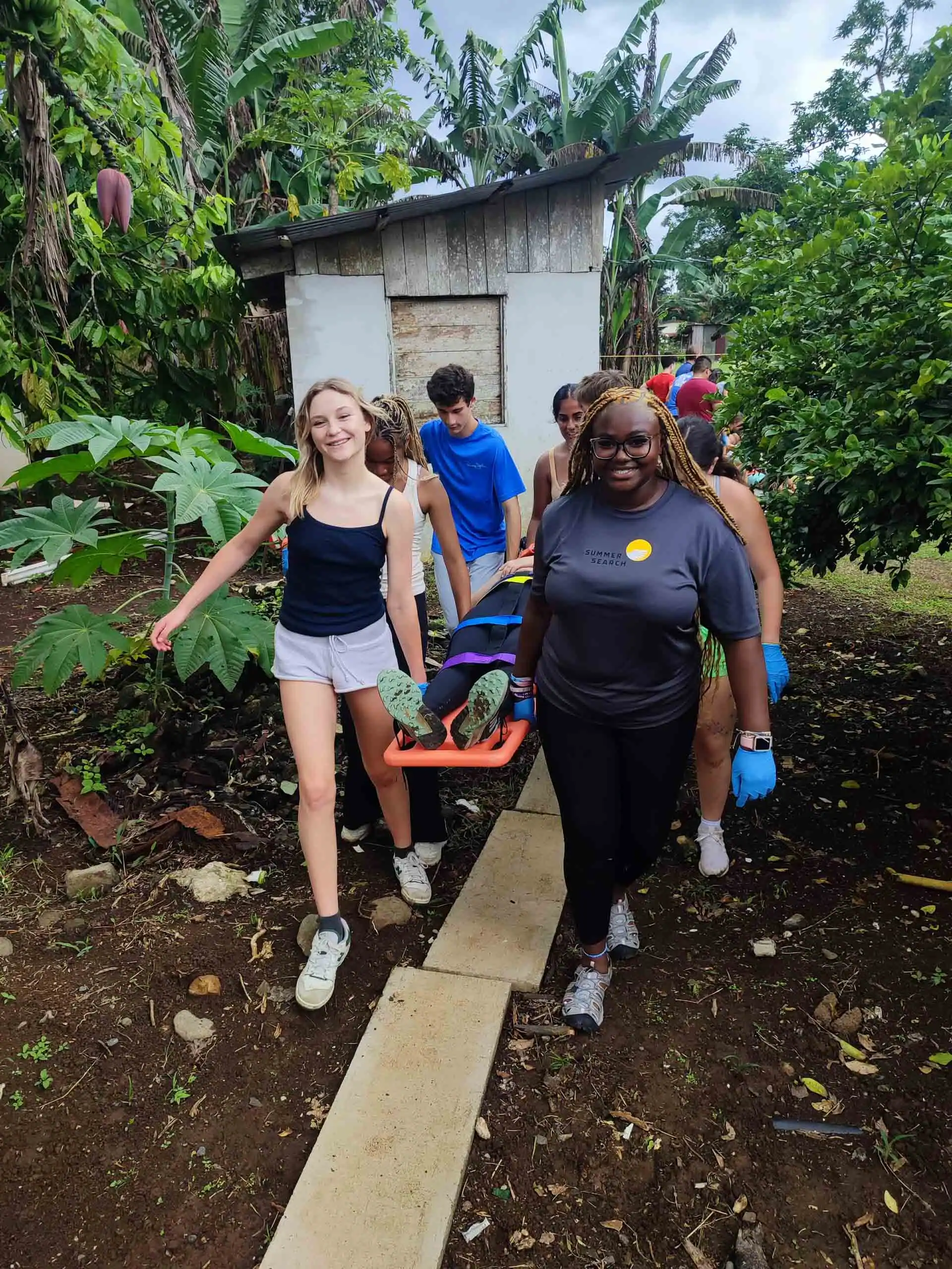 A group of students and medical professionals, during a medical study abroad program in Costa Rica, carrying a person on a stretcher along a garden path, with greenery and a small shed in the background.