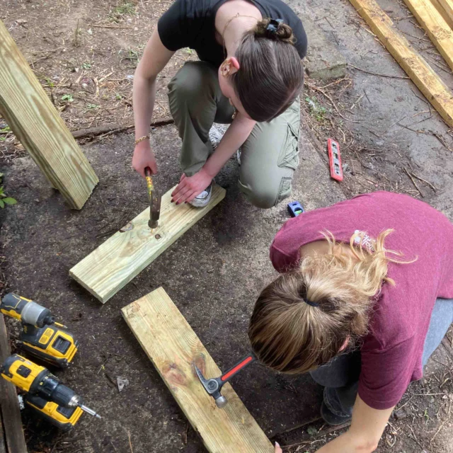 Two people crouch on the ground, assembling wooden beams with drills and a hammer. Construction tools are scattered around them.