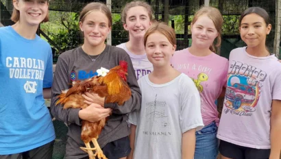 Six young people stand together, one holding a chicken, in front of a coop.