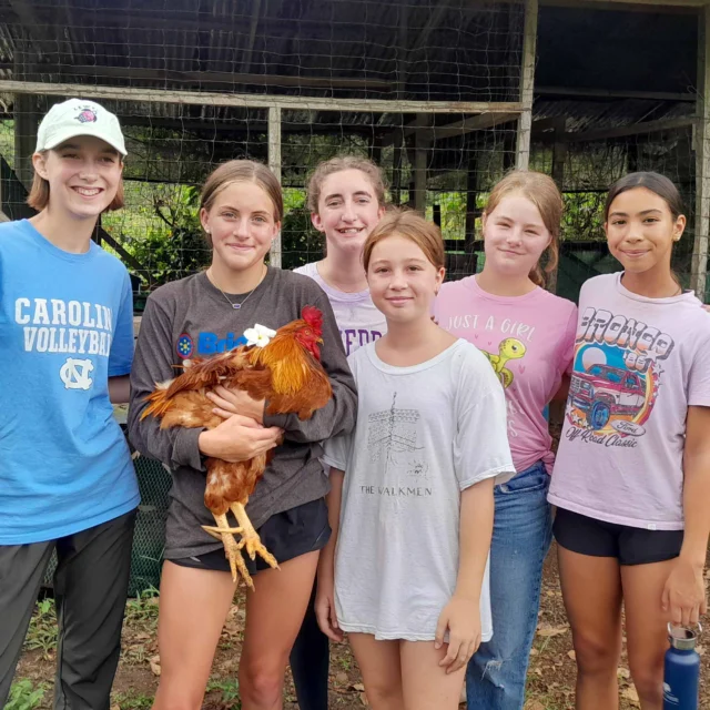 Six young people stand together, one holding a chicken, in front of a coop.