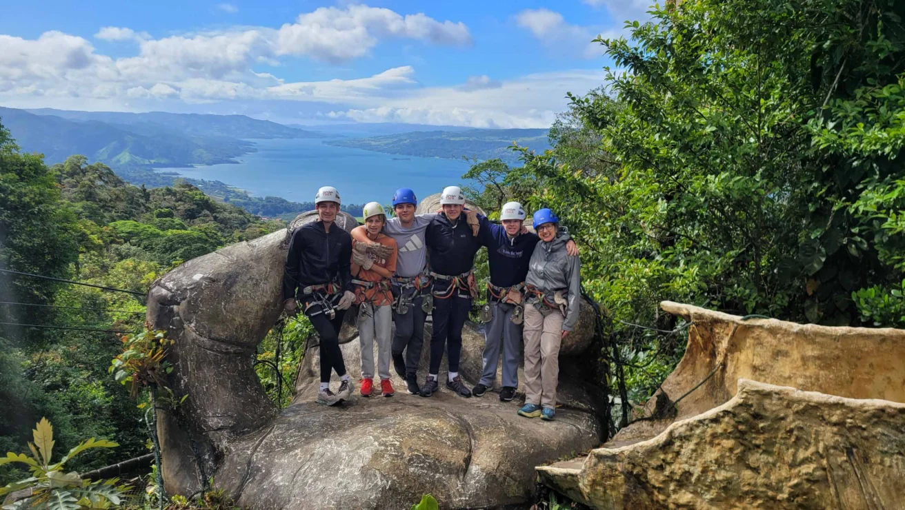 A group of six people wearing helmets and safety gear stand on a large rock, with a scenic view of a lake and mountains in the background.