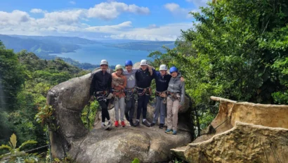 A group of six people wearing helmets and safety gear stand on a large rock, with a scenic view of a lake and mountains in the background.