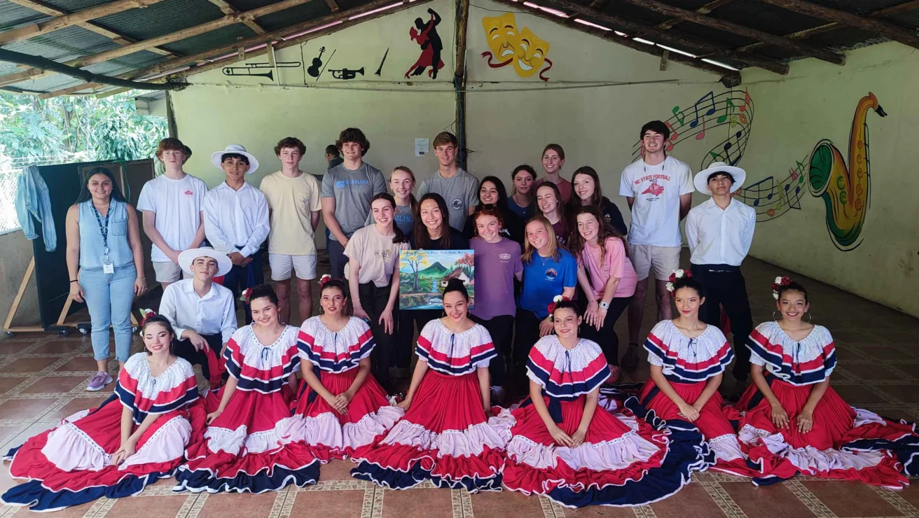 A group of people pose indoors, with some wearing traditional red, white, and blue dresses. The backdrop features musical and theatrical artwork.